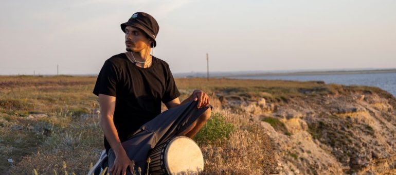 A man in a bucket hat sits with a drum on a grassy cliffside, gazing into the distance at sunset.